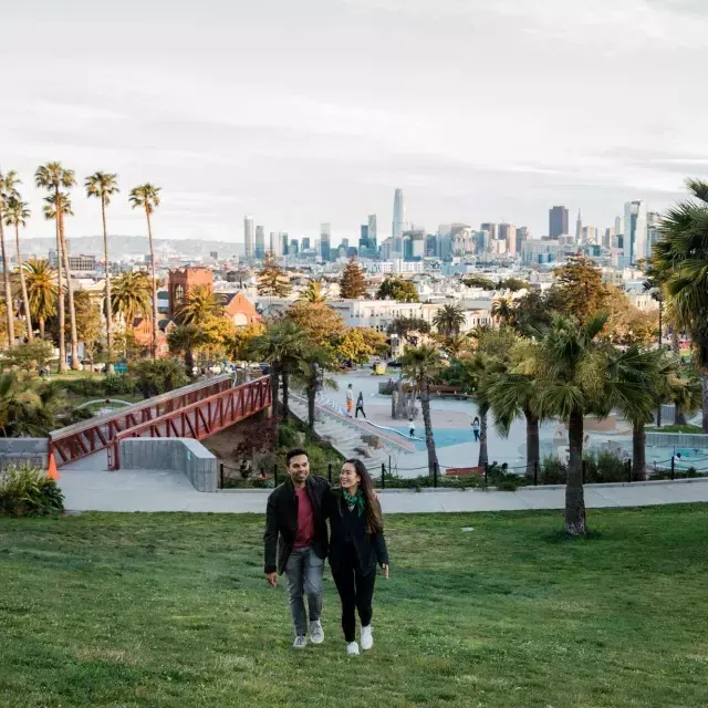 Un couple se dirige vers la caméra avec Dolores Park et la Skyline de San Francisco derrière eux.